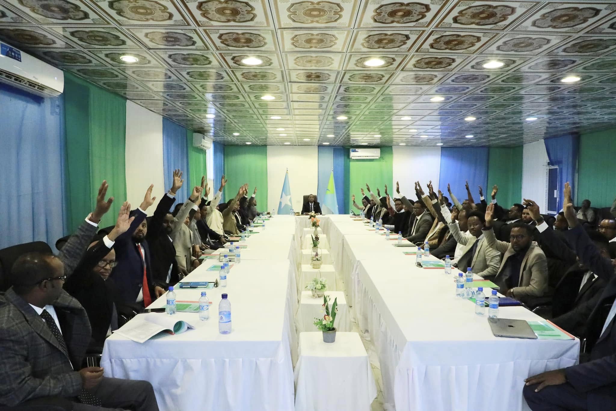 A group of officials from the Hirshabelle Council of Ministers participating in a ribbon-cutting ceremony, surrounded by flowers and an archway, during an event in the Balcad district. Photographers are present documenting the moment.