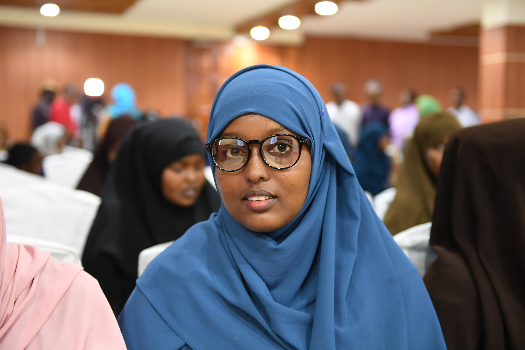 A Somali woman wearing a blue hijab and glasses attends an event, seated among a group of attendees, in a brightly lit hall.