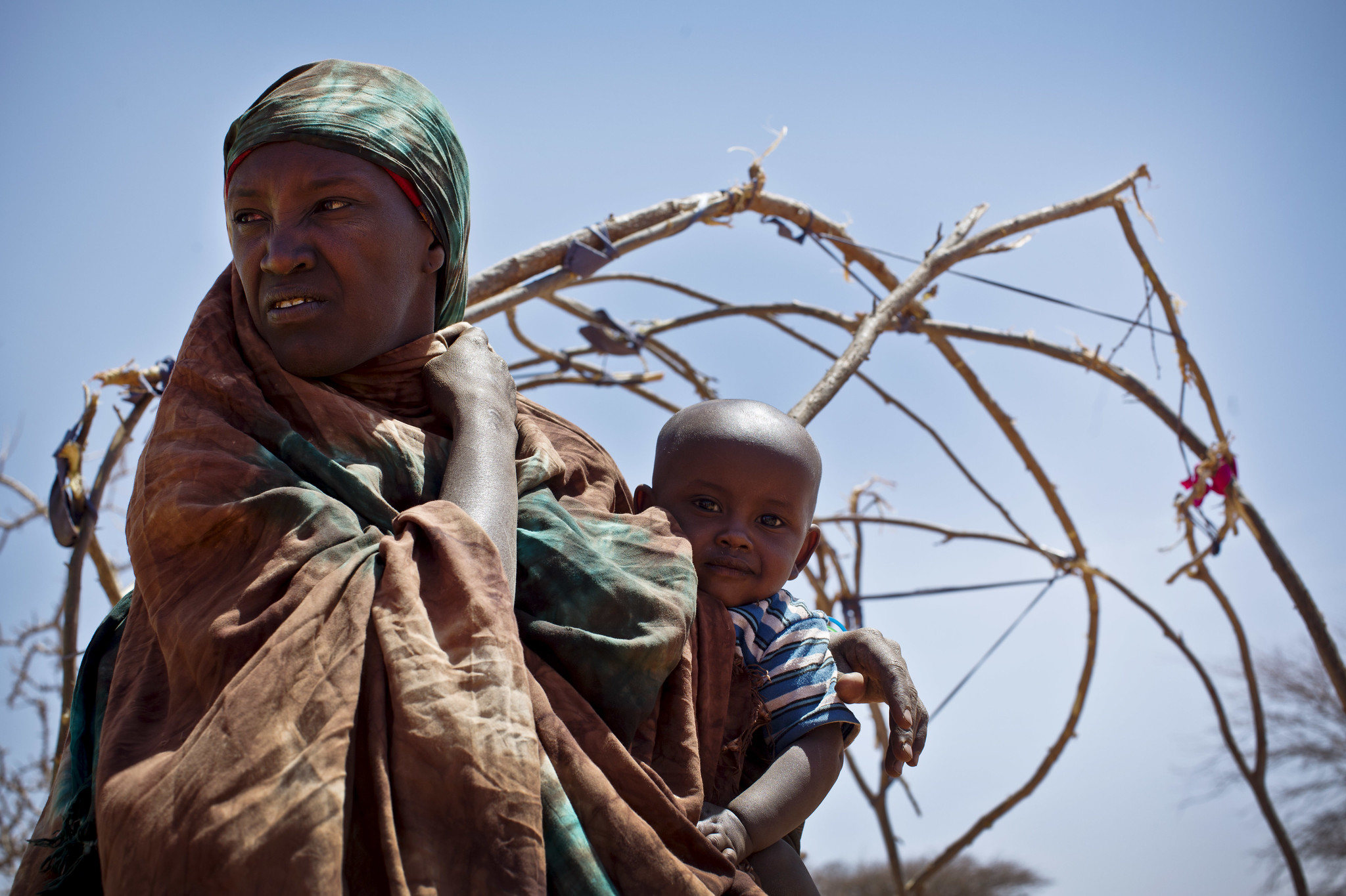 A Somali mother holds her child while standing in front of an unfinished makeshift shelter constructed with branches, reflecting the impact of severe drought near Ainabo, Somalia.