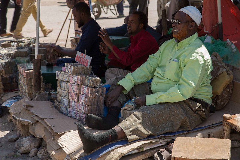 Photo taken in 2015 in Hargeisa by Dawan TV. It depicts two men sitting by a market, one as a money changer and the other as a customer. They are seated on makeshift chairs with other people walking by in the background