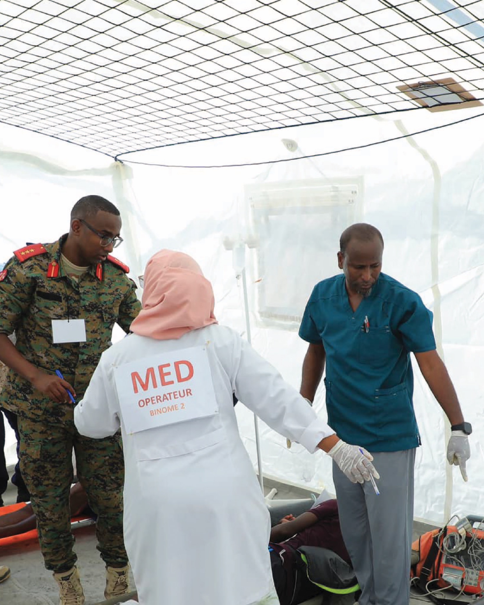 Medical and military personnel participate in a maritime rescue exercise named "Coordination" in Djibouti. A female healthcare worker in a white coat labeled "MED OPERATEUR," a military officer in camouflage uniform, and a male doctor in blue scrubs are seen attending to a simulated patient inside a temporary medical tent.