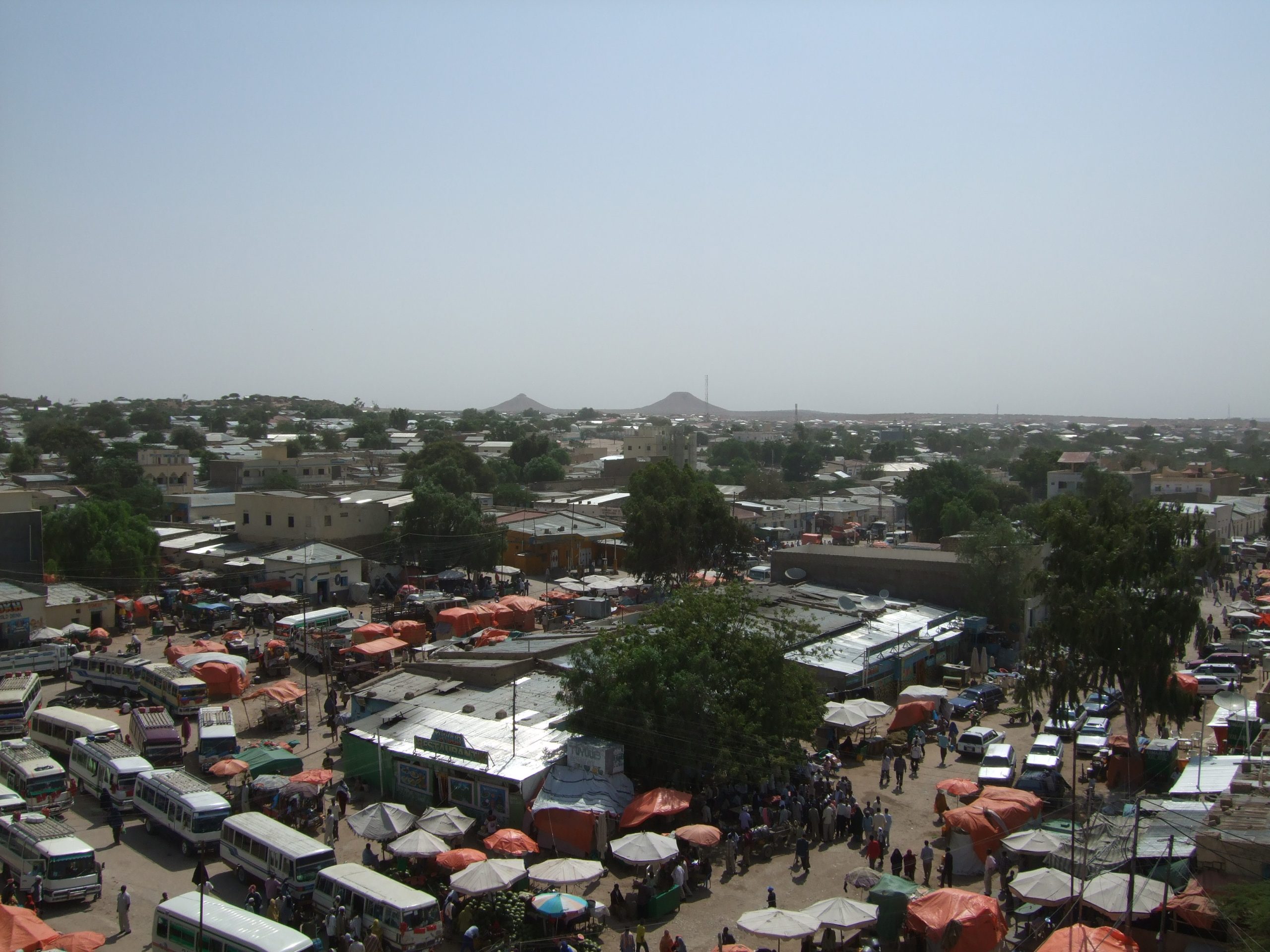 The picture was taken in 2008 and shows a busy market in Hargeisa. People, vehicles, and market stalls can be seen. In the background, there are multi-story buildings and the city's hills.