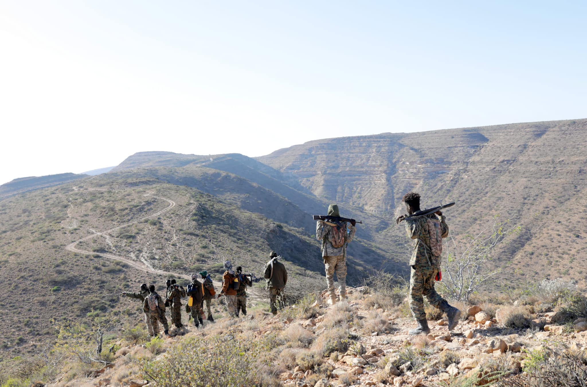 Armed soldiers in camouflage uniforms patrol the Cal Miskaad Mountains in Somalia. Some carry weapons on their shoulders while others appear to be scouting the rugged terrain.