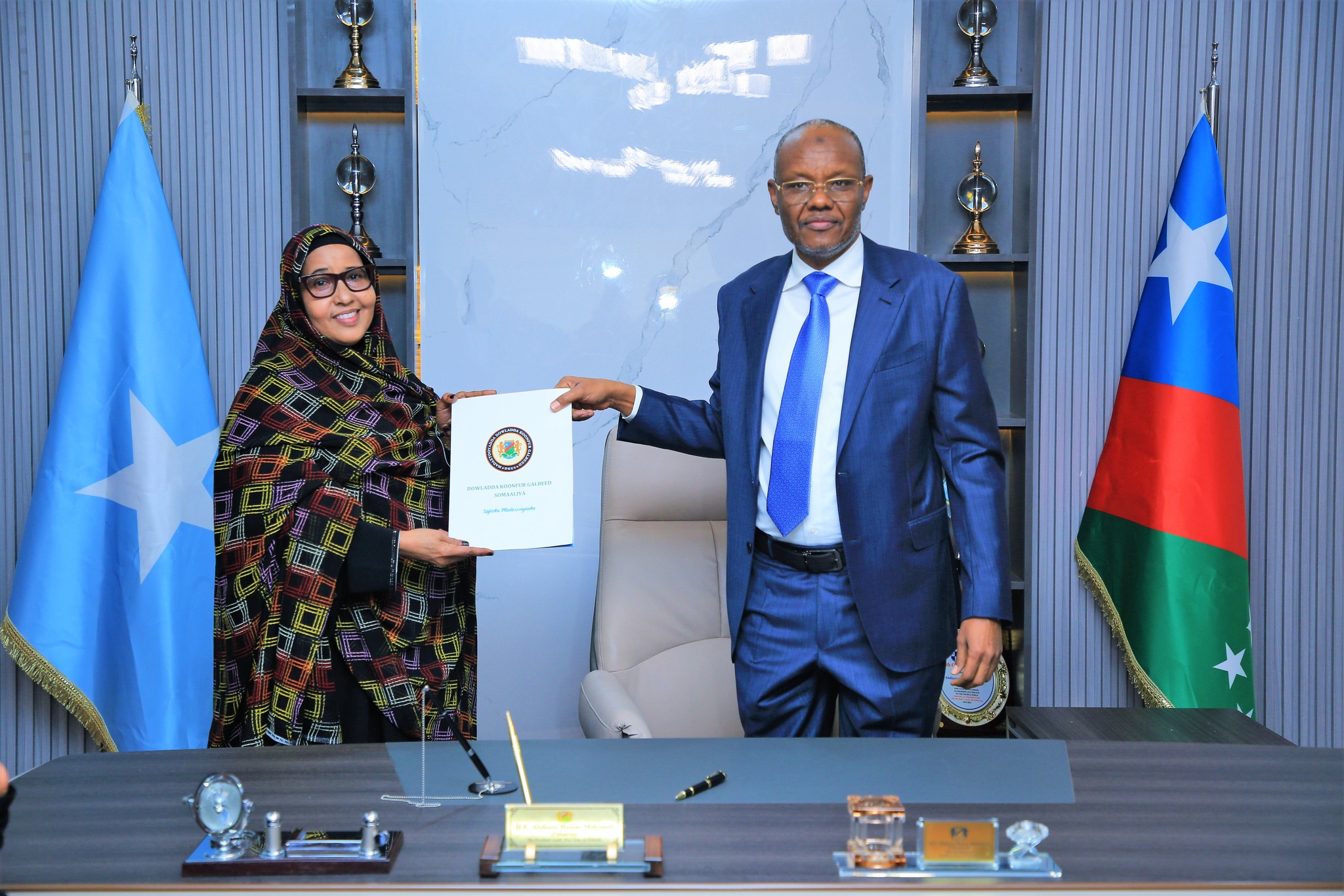 Southwest State president and the minister of women smiling as they present a document in an official setting, with the flags of Somalia and the Southwest State in the background