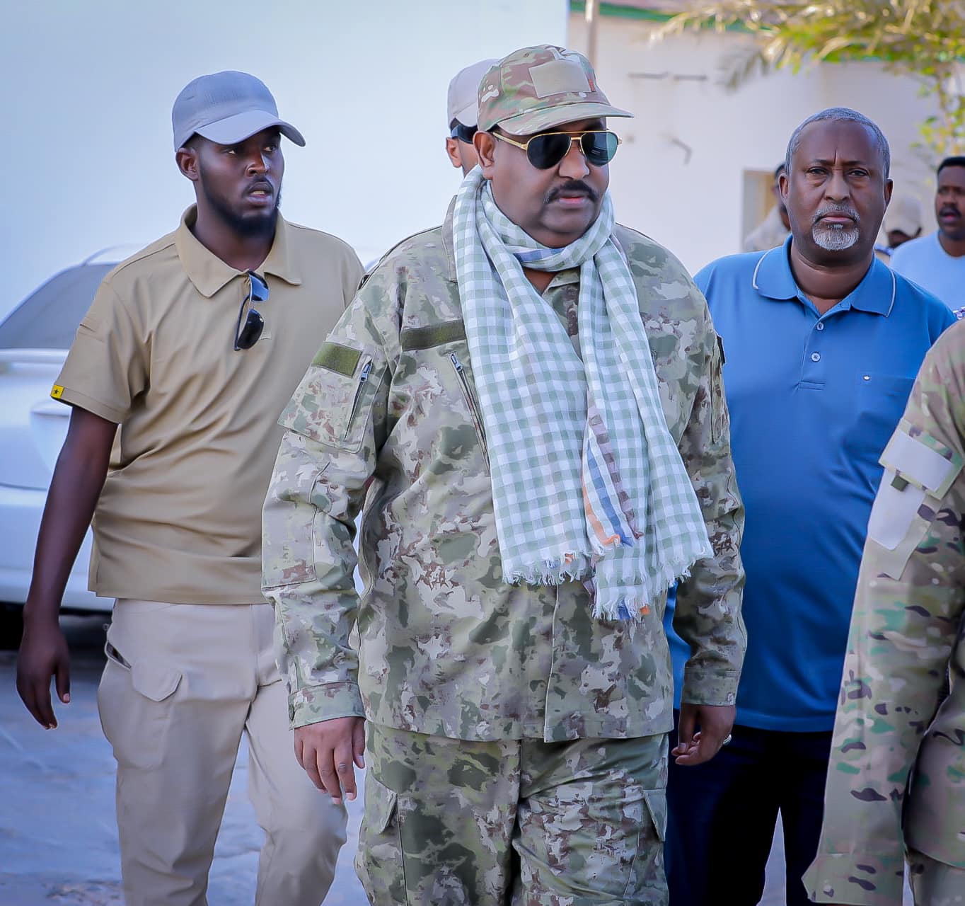 A high-ranking Somali official in military fatigues and sunglasses, accompanied by security personnel. The official is wearing a camouflage uniform and a checkered scarf.