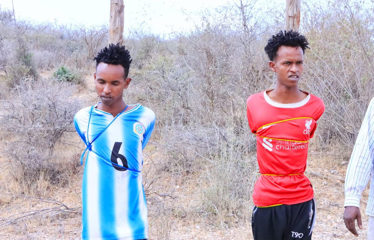 Two men, Mukhtaar Mohamed Nur and Nur Mohamed Hassan, are tied against wooden poles in an arid, bushy area, awaiting execution by firing squad. One is wearing a blue and white jersey with the number 6, and the other is wearing a red Liverpool football jersey.