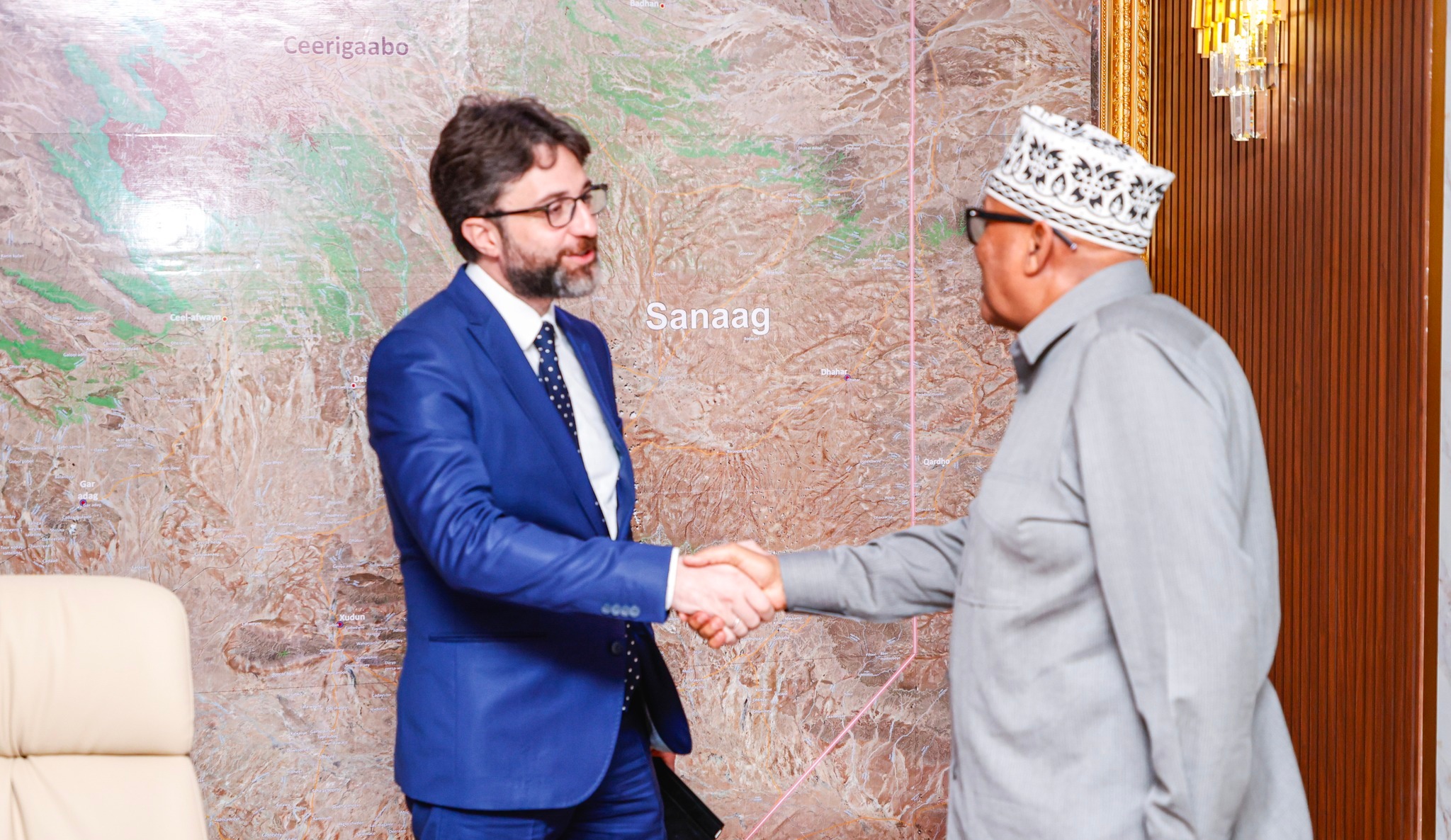 Somaliland President Cabdiraxmaan Maxamed Cabdillaahi (Cirro) shakes hands with Turkey’s Ambassador to Somalia in front of a large map of Sanaag region. The president is dressed in traditional Somali attire with a kufi cap, while the ambassador wears a blue suit and tie.