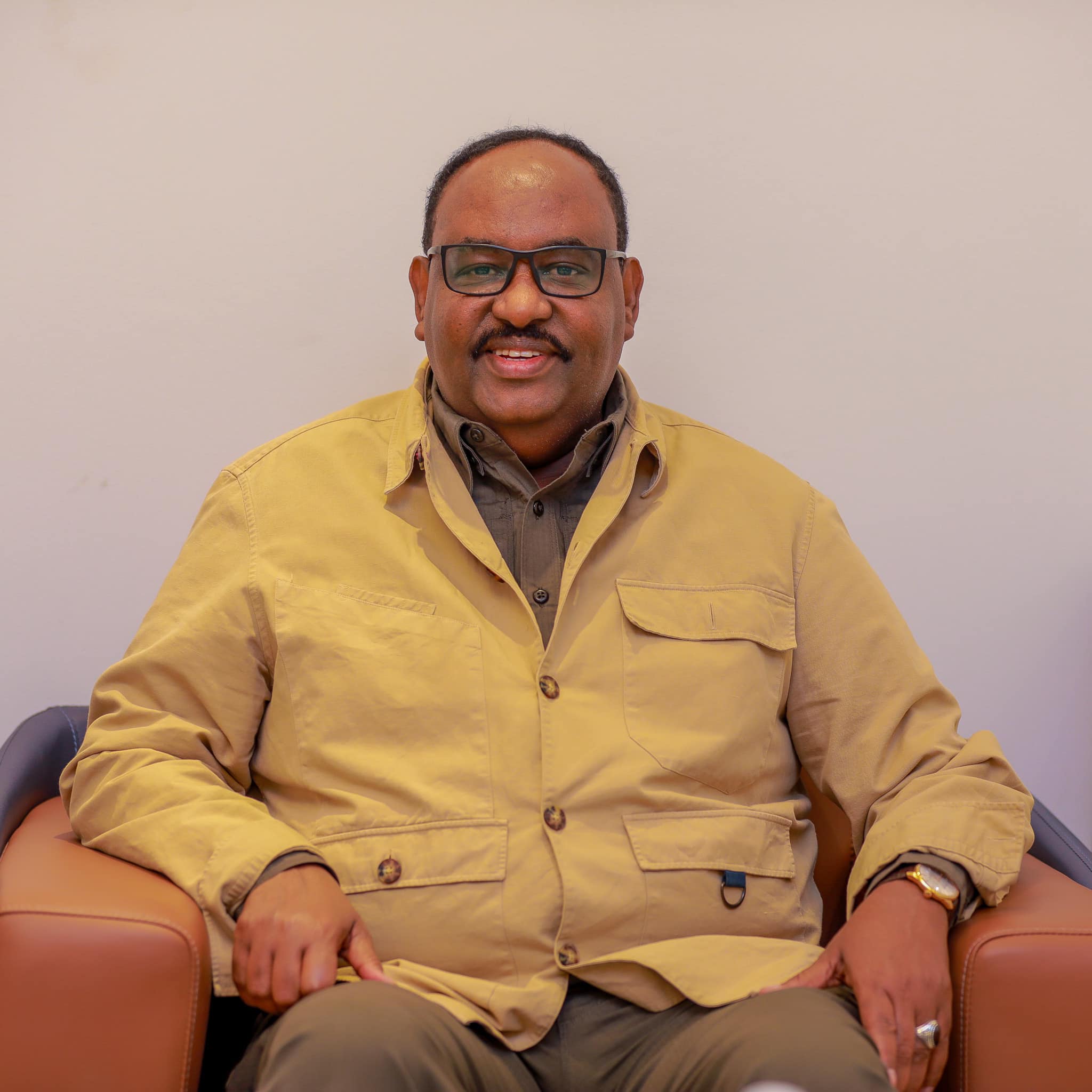 A portrait of Said Abdullahi Deni, President of Puntland, wearing a beige jacket and dark brown shirt, seated in a relaxed posture.