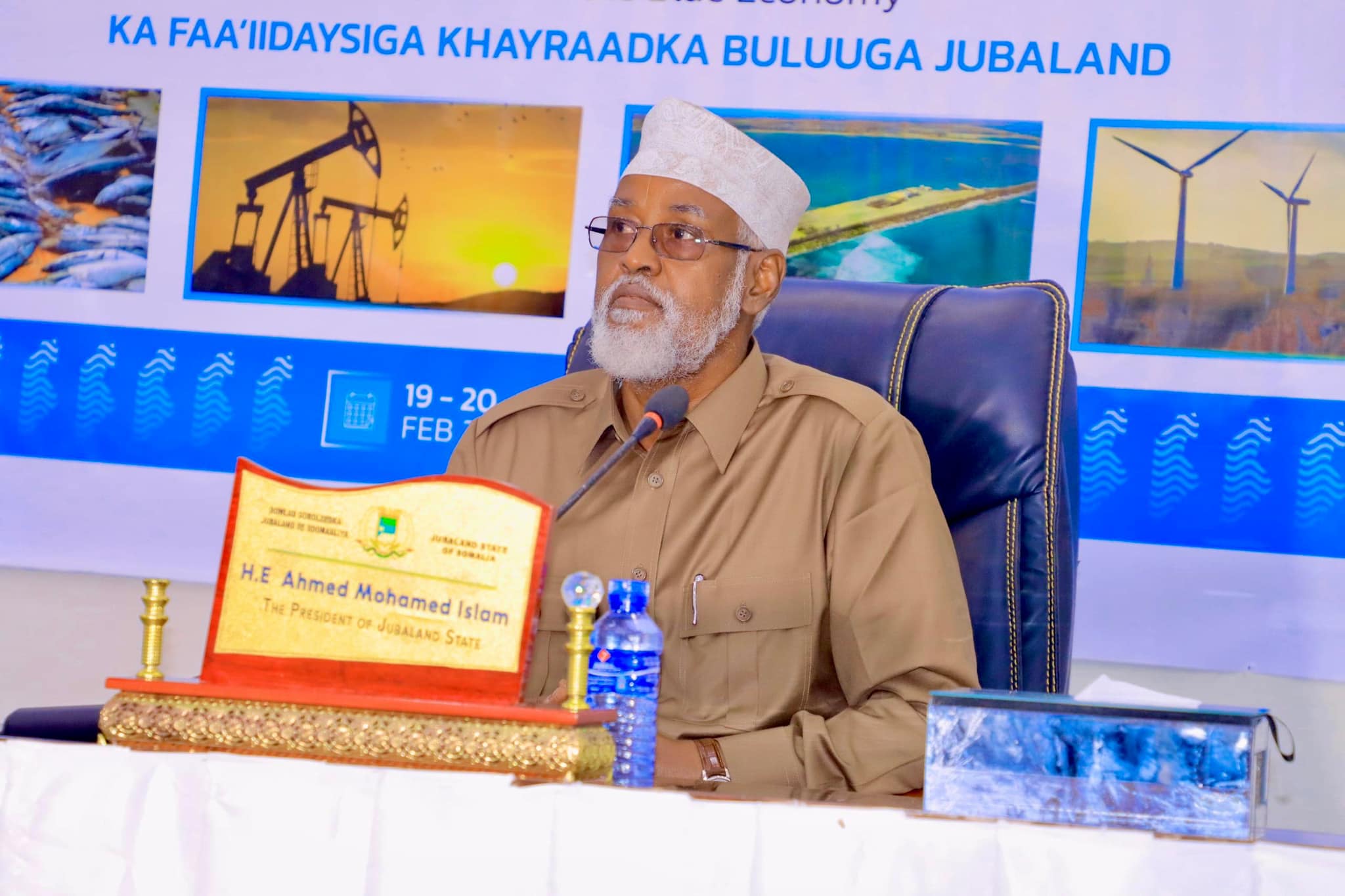 Ahmed Mohamed Islam, President of Jubaland State, seated at a conference table during the "Blue Economy" forum focused on utilizing Jubaland's marine resources. A backdrop displays images of fish, oil rigs, wind turbines, and coastal views.