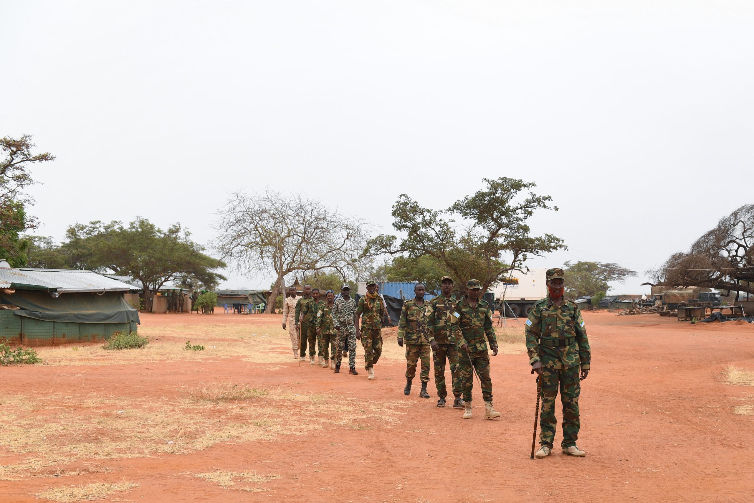 A group of Somali National Army (SNA) soldiers in camouflage uniforms walking in formation at a military camp in a dry, rural environment. The leading soldier holds a stick while the others follow behind.