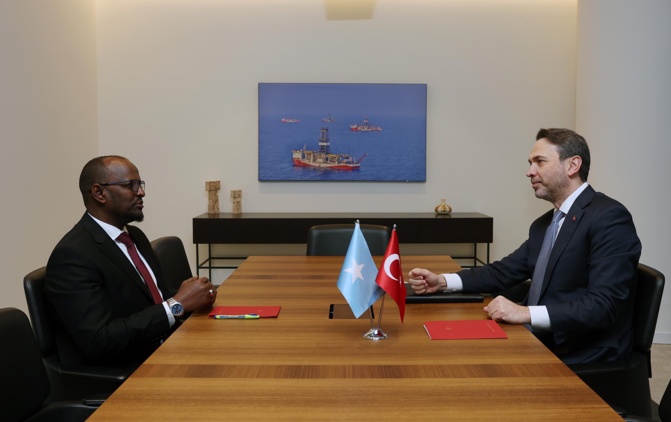 Two officials, one representing Somalia and the other representing Turkey, sit across from each other at a conference table with Somali and Turkish flags in the center. They are engaged in a diplomatic discussion in a professional setting. A framed image of offshore oil drilling platforms is displayed on the wall behind them.