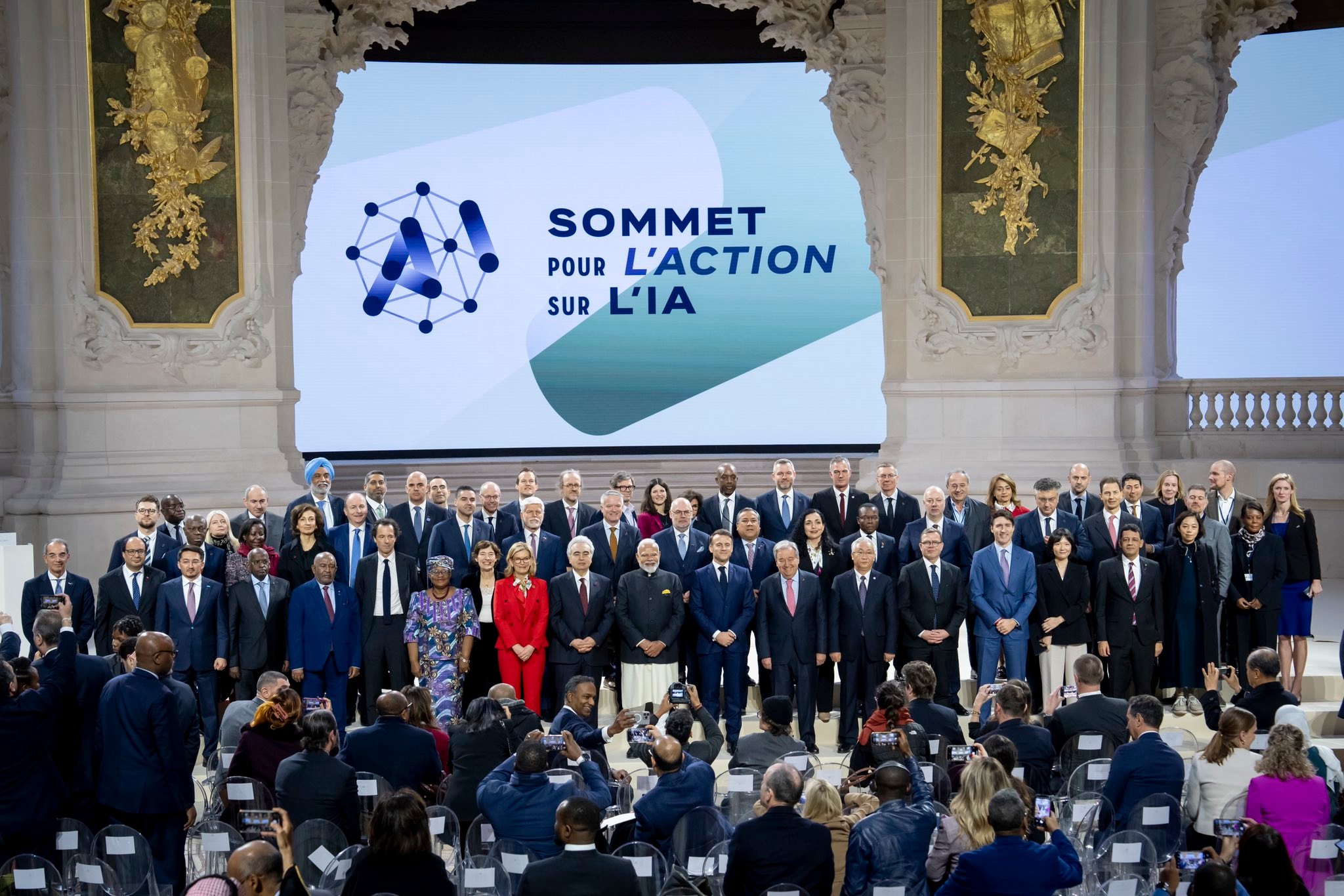 A large group of world leaders, policymakers, and experts gather for a group photo at the AI Action Summit (Sommet Pour L'Action Sur L'IA). The event is taking place in an elegant conference hall with a grand stage displaying the summit’s branding.