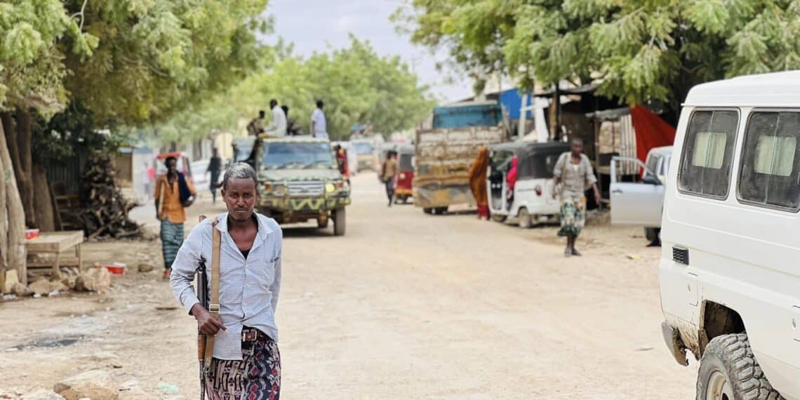 A member of the Ma'awiisley militia with a rifle slung over his shoulder walking down a street with military vehicles and civilians in the background.