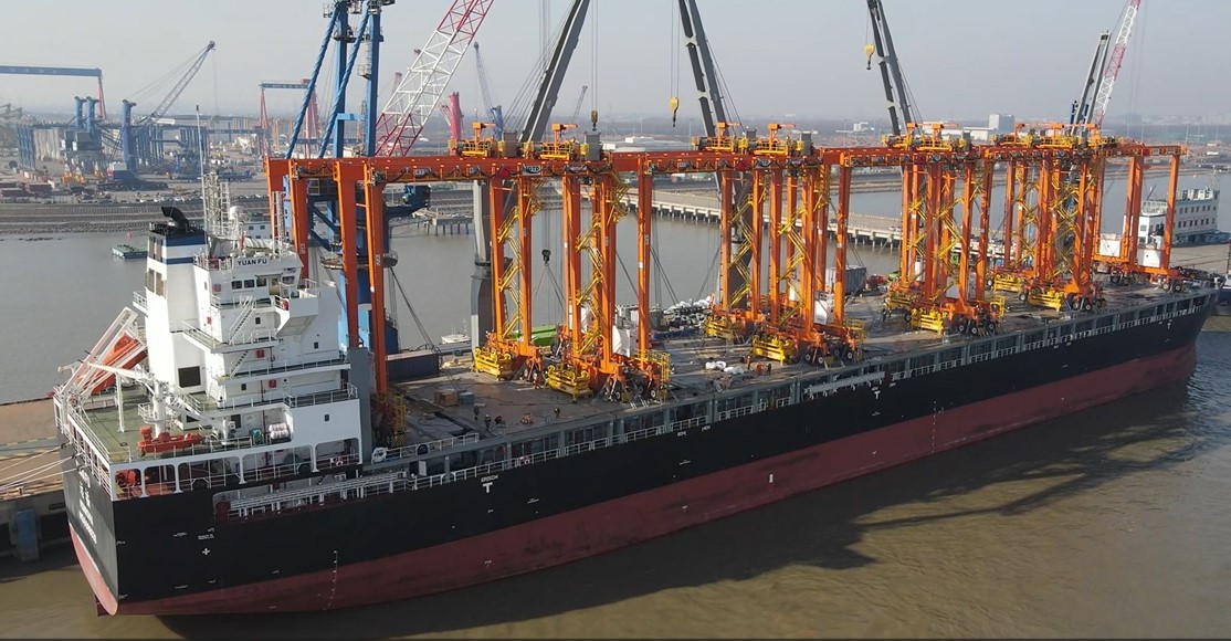 A large cargo ship docked at a port, carrying multiple bright orange rubber-tired gantry (RTG) cranes. Several cranes and port infrastructure are visible in the background.