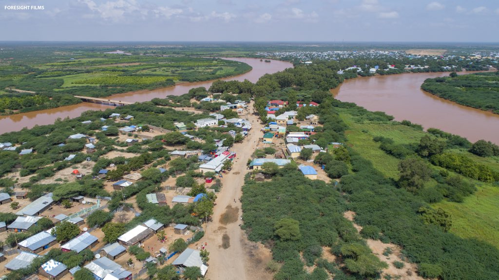 An aerial view of Luuq, a town in Somalia, situated along the Juba River. The image shows houses with metal roofs, green vegetation, and a bridge connecting different parts of the town.