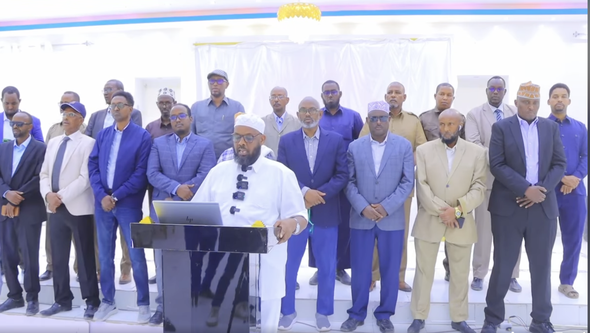 A group of SSC-Khaatumo lawmakers stand behind a speaker at a press conference. The speaker, dressed in white traditional attire with a white cap and glasses, is addressing the audience from a podium with a laptop.