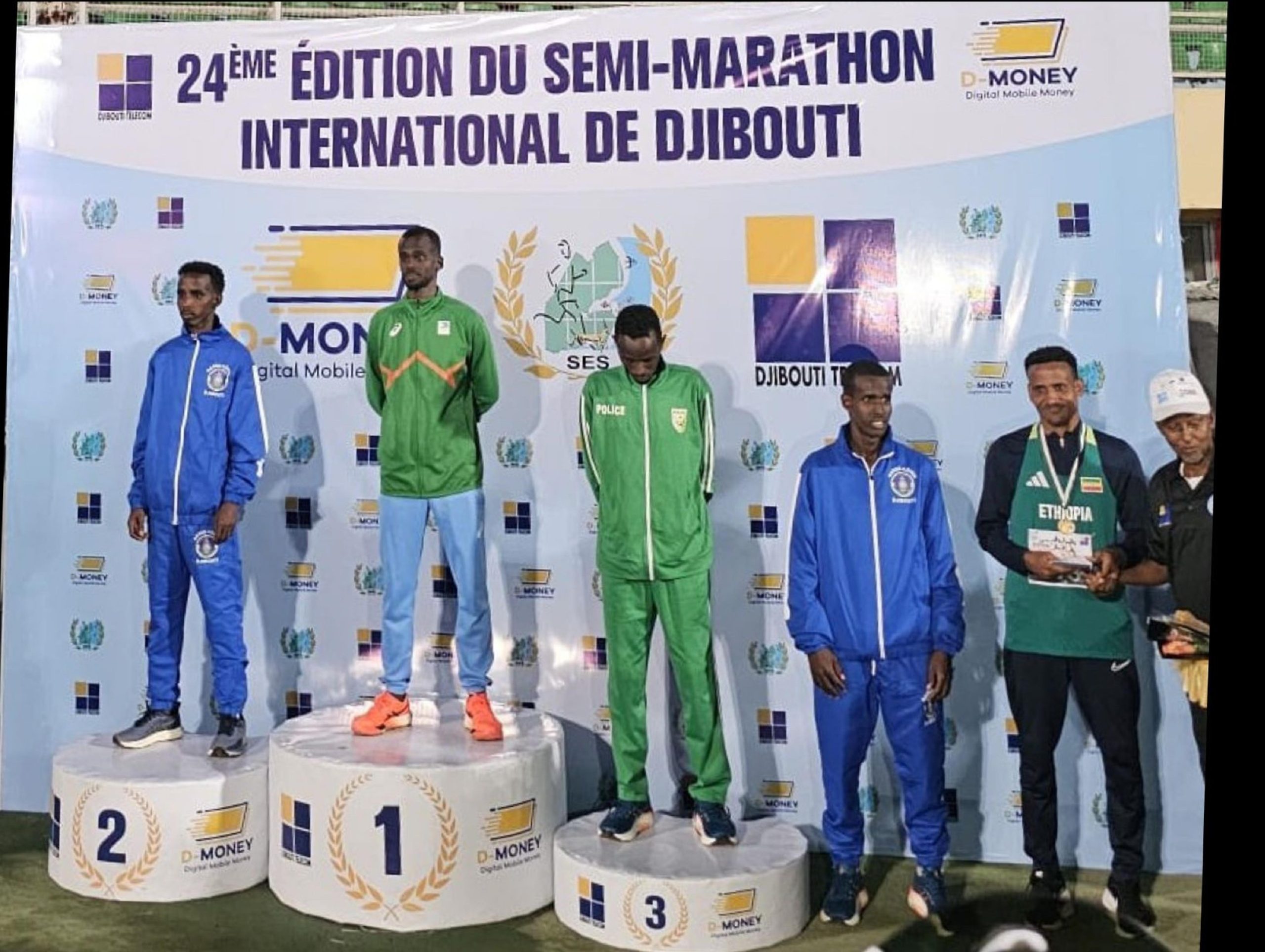 Athletes stand on the winners' podium at the 24th edition of the Djibouti International Semi-Marathon. The first-place winner wears a green tracksuit, the second and third-place finishers wear blue tracksuits, and another athlete in an Ethiopian uniform is holding a trophy. A banner with event sponsors and logos is visible in the background.