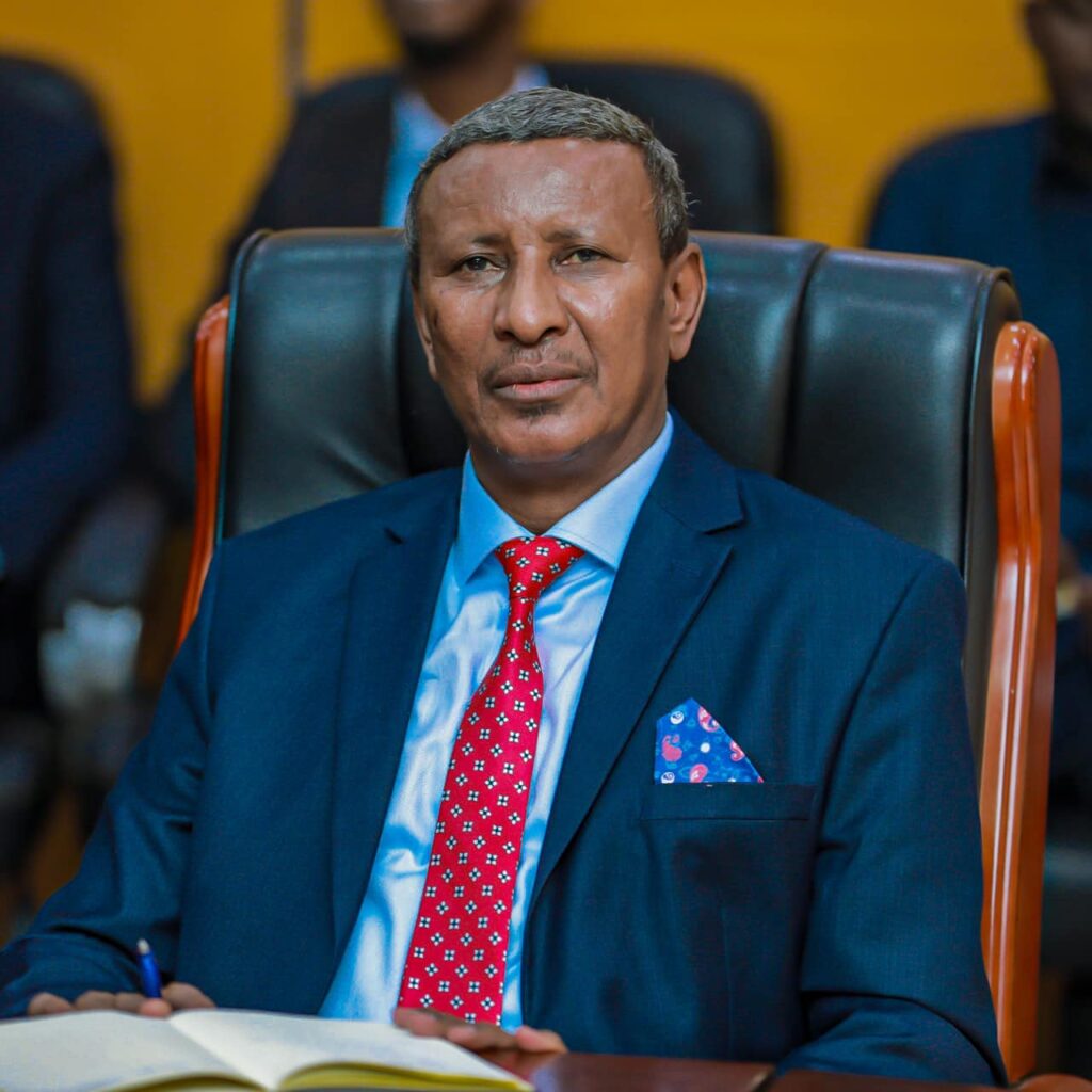 Puntland State Minister of Information Maxamuud Caydiid Dirir sitting attentively in a meeting, dressed in a blue suit with a red tie.