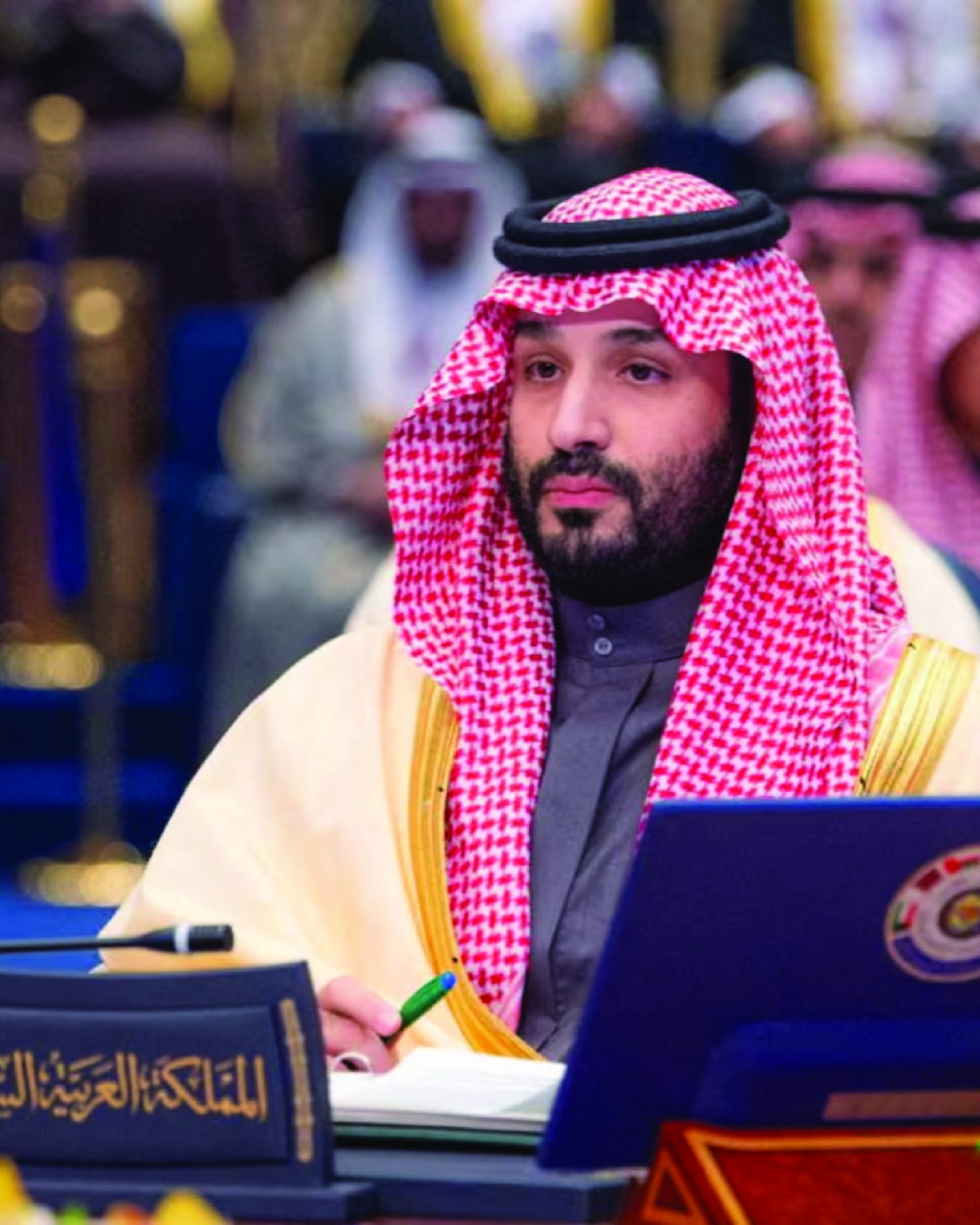 Crown Prince Mohammed bin Salman of Saudi Arabia, dressed in traditional attire with a red and white checkered ghutra and a black agal, is seated at an international summit. He holds a pen and appears attentive, with a nameplate reading "Kingdom of Saudi Arabia" in front of him.