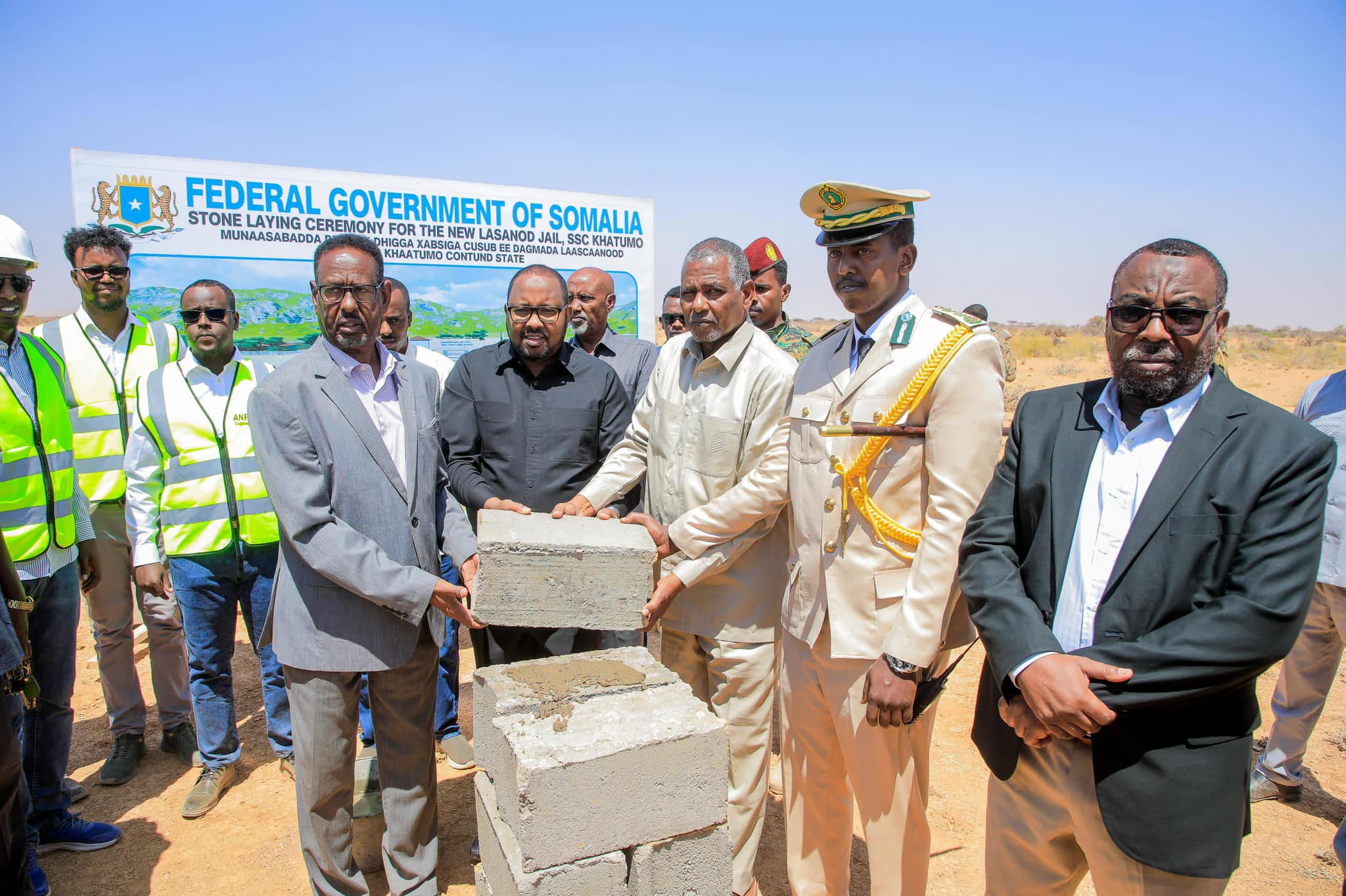 Officials and security personnel participate in the foundation stone laying ceremony for the new Lasanod jail, SSC-Khaatumo, funded by the Federal Government of Somalia. They are dressed in formal and security attire, standing next to construction blocks at the event site under a clear blue sky.