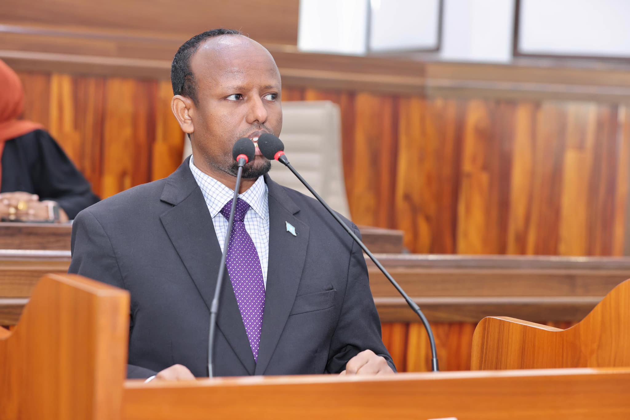 A Somali government official, identified as Minister of Health Ali Haji, speaks at a podium inside a parliamentary hall, addressing the audience.