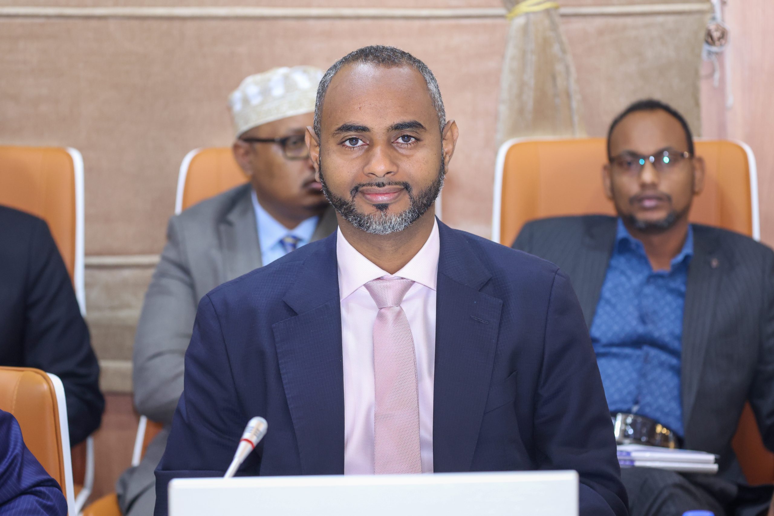 Somalia’s Minister of Defense, Cabdiqaadir Maxamed Nuur, seated at an important meeting, wearing a navy blue suit and pink tie.