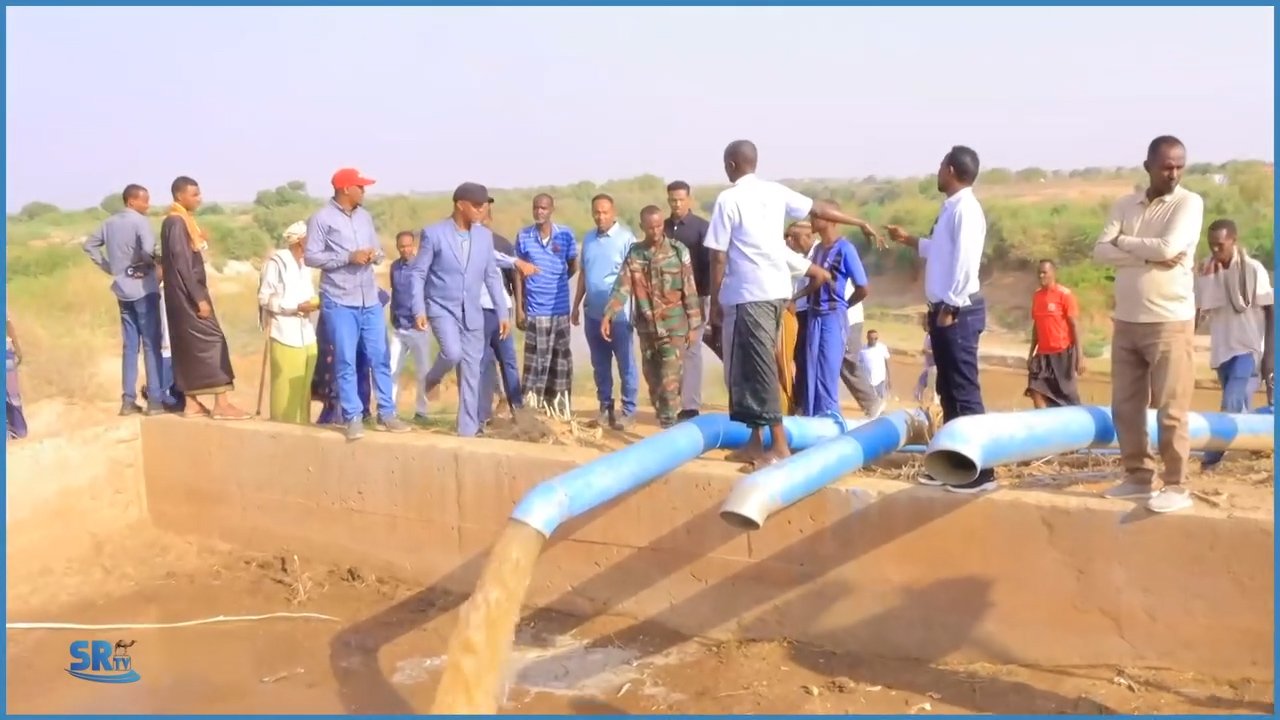 Group of people, including officials and farmers, inspecting a modern irrigation system with large blue pipes pumping water from the Shabelle River in a rural setting.