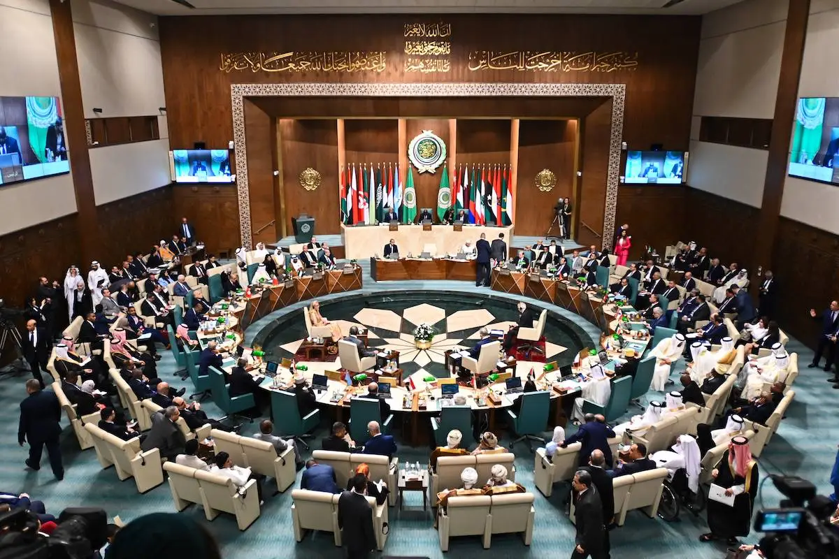 A high-level diplomatic meeting at the Arab League headquarters, featuring a circular arrangement of officials representing various Arab nations. Delegates, many in traditional attire, are seated around a large conference table adorned with flags, microphones, and documents.