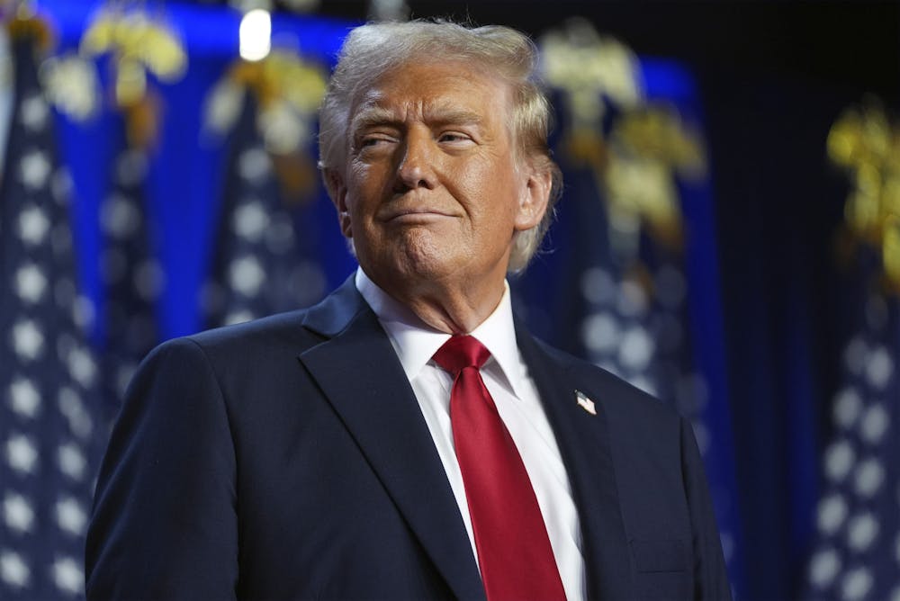 Former U.S. President Donald Trump standing in front of American flags, wearing a dark suit and red tie.