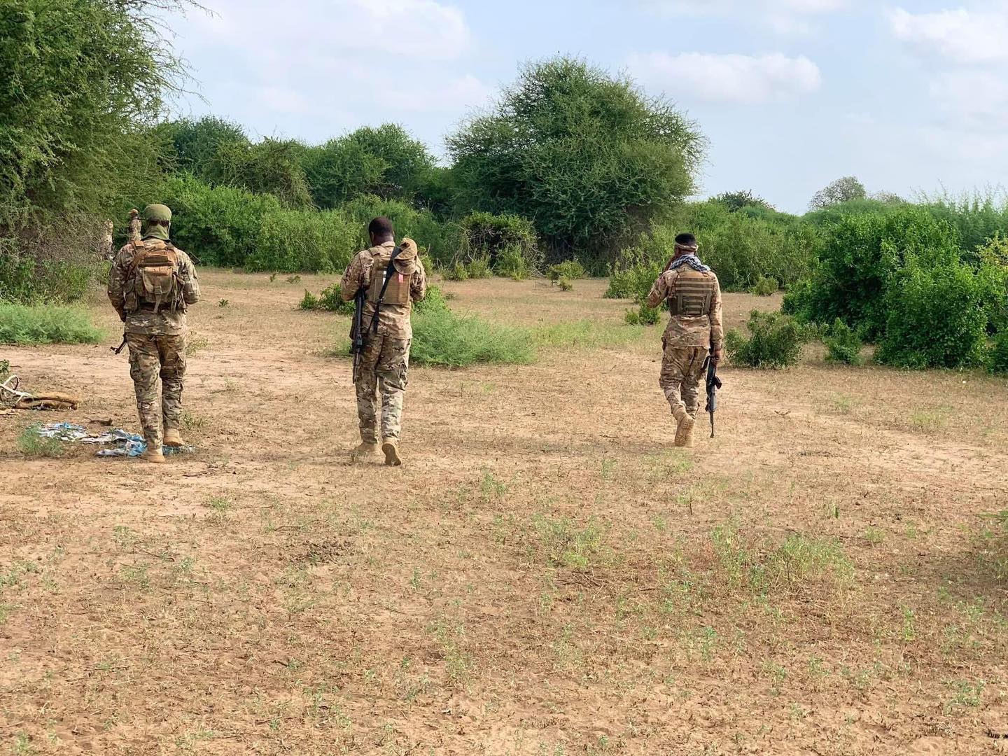 Three soldiers in camouflage uniforms walking away through a dry, sparsely vegetated field with green bushes under a partly cloudy sky. Caption: