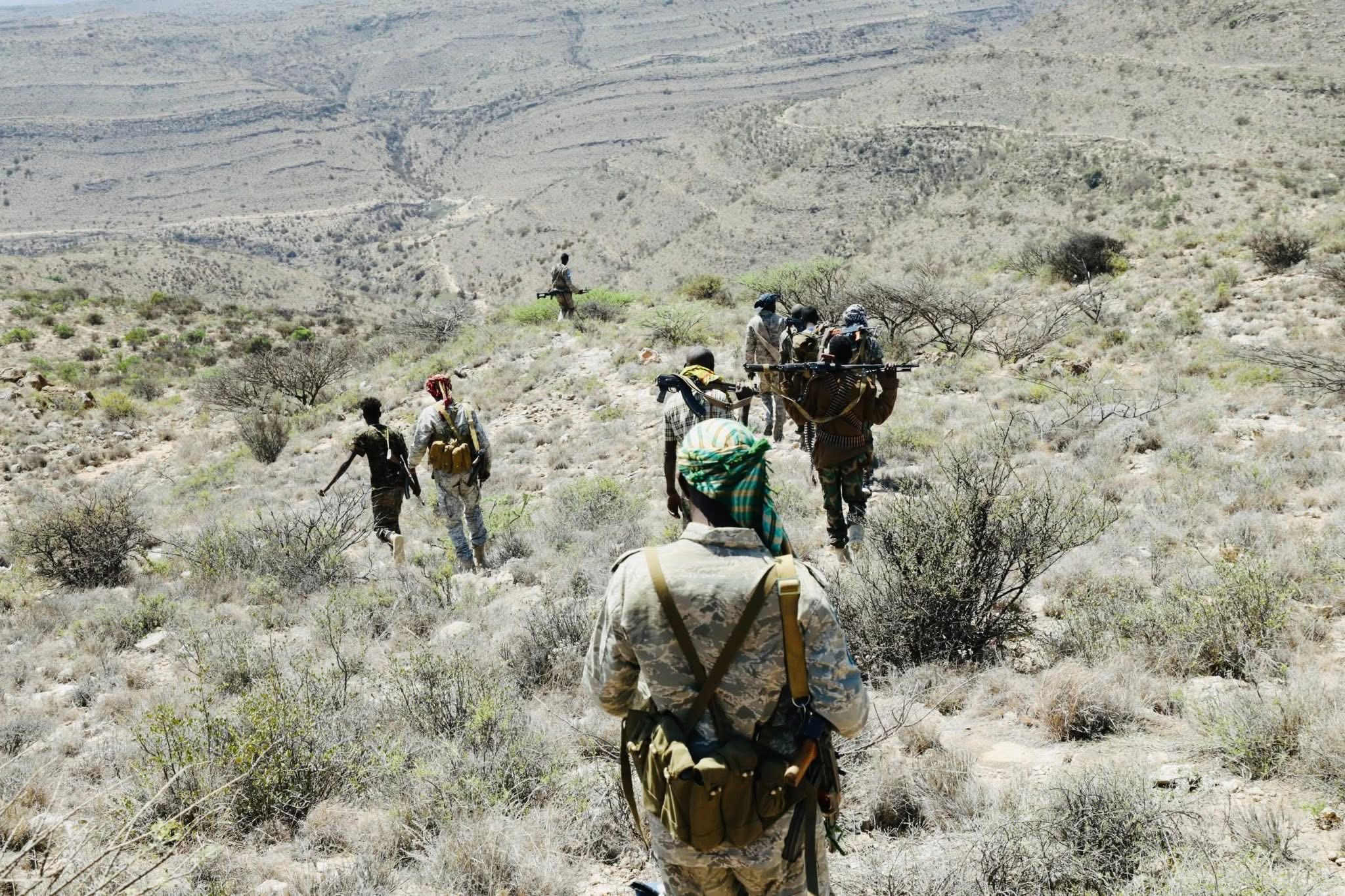 Armed soldiers trekking through a dry mountainous area with sparse trees and shrubs.