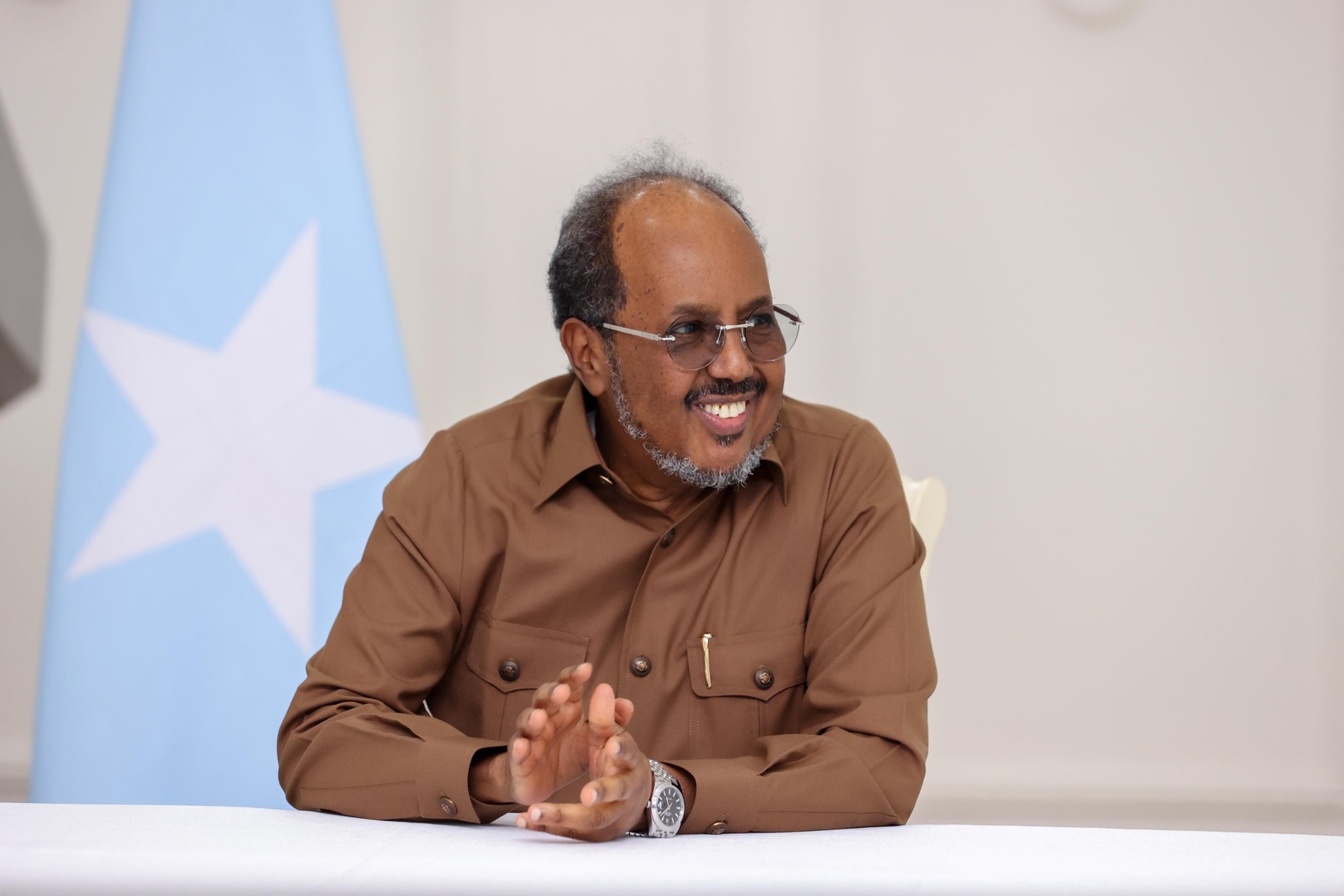 Somali leader smiling while seated at a table with the national flag in the background.